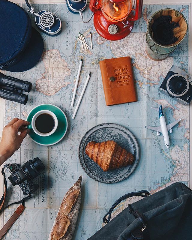 Photo of coffee, camera, baked goods, travel journals, headphones, travel bag, lantern, matches and mini airplanes over a map of the world.