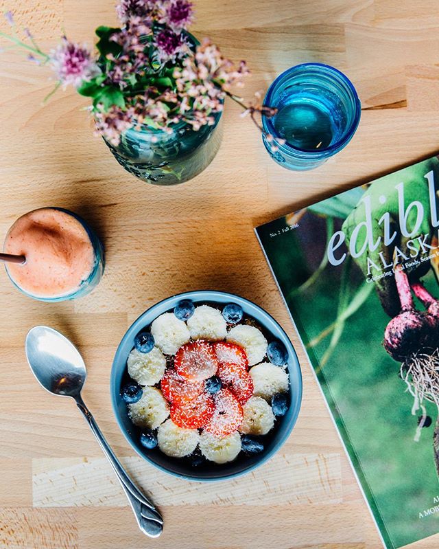 A photo of an acai bowl with spoon, smoothie, plant, glass of water and magazine on a table.