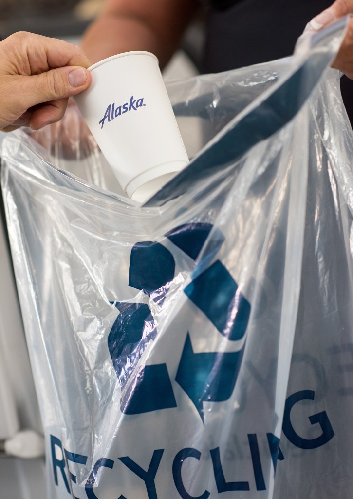 A photo of a customer throwing an Alaska coffee cup in a recycling bag held by an Alaska Airlines flight attendant onboard an Alaska Airlines aircraft.