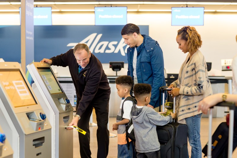 A photo of a male Alaska Airlines agent helping print bag tags at a Self-Tag station for a family of 4 traveling together.