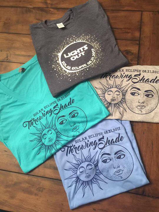 Photo of four shirts laying on table. Design features an illustration of a moon glaring at a sun with the words "Throwing Shade" above.