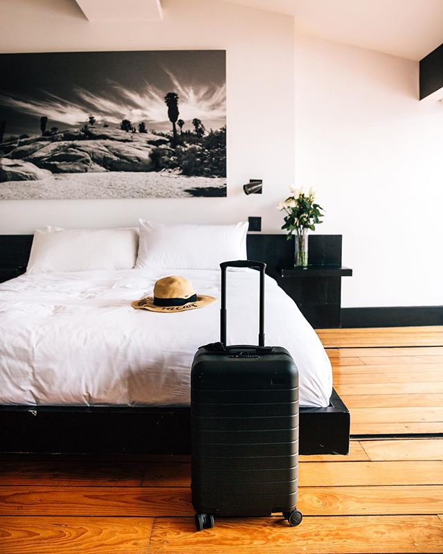 This is a photo of a hotel room with wood floors, a large bed, a suitcase and a hat sitting on the bed.
