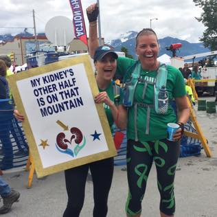 This is a photo of Jenny Stansel holding a sign that says "My Kidney's Other Half is on that Mountain!" while standing next to Jodi Harskamp at the finish line of a race.