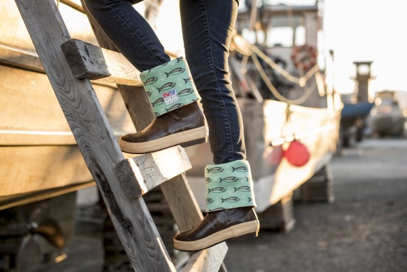 This is a closeup photo of a girl climbing up into a boat wearing Salmon Sisters branded boots. The boots are brown with a blue salmon and hook design.
