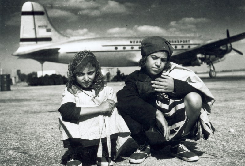 This is a historical, black and white photo of two young Yemenite Jews sitting on the tarmac with a DC-4 aircraft behind them.