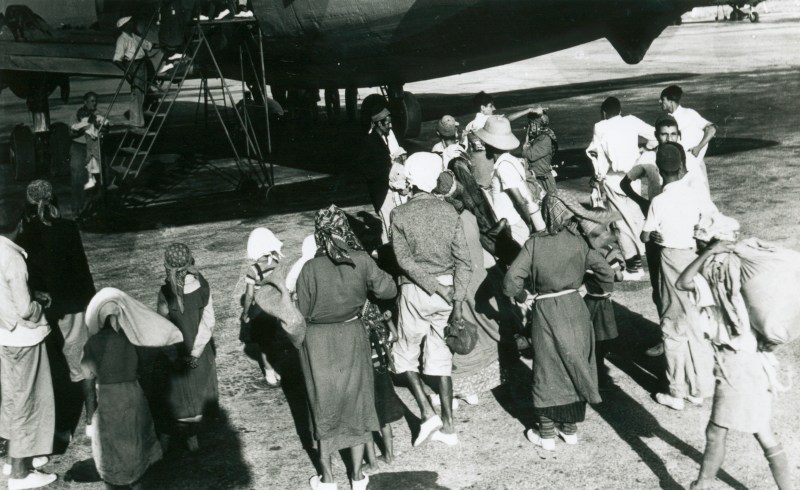 This is a historical, black and white photo of a group of about 15 Yemenite Jews gathering beside the entrance of an airplane.