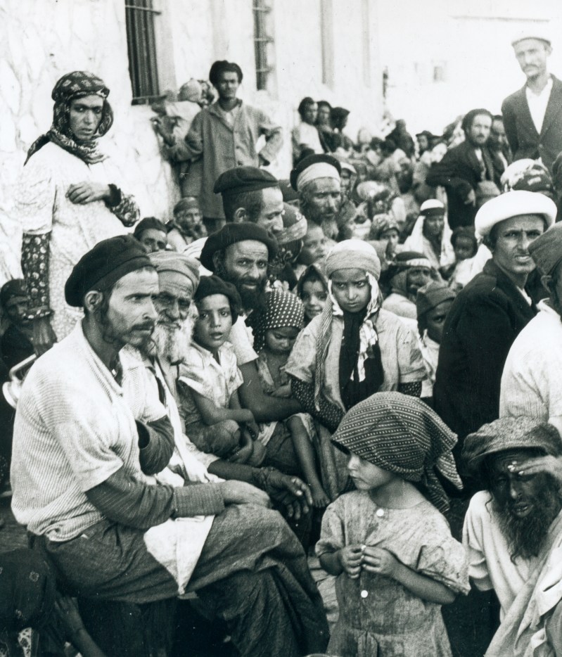 This is a historical photo in black and white print of a group of about 50 Yemenite Jews awaiting transport on the streets of Aden.